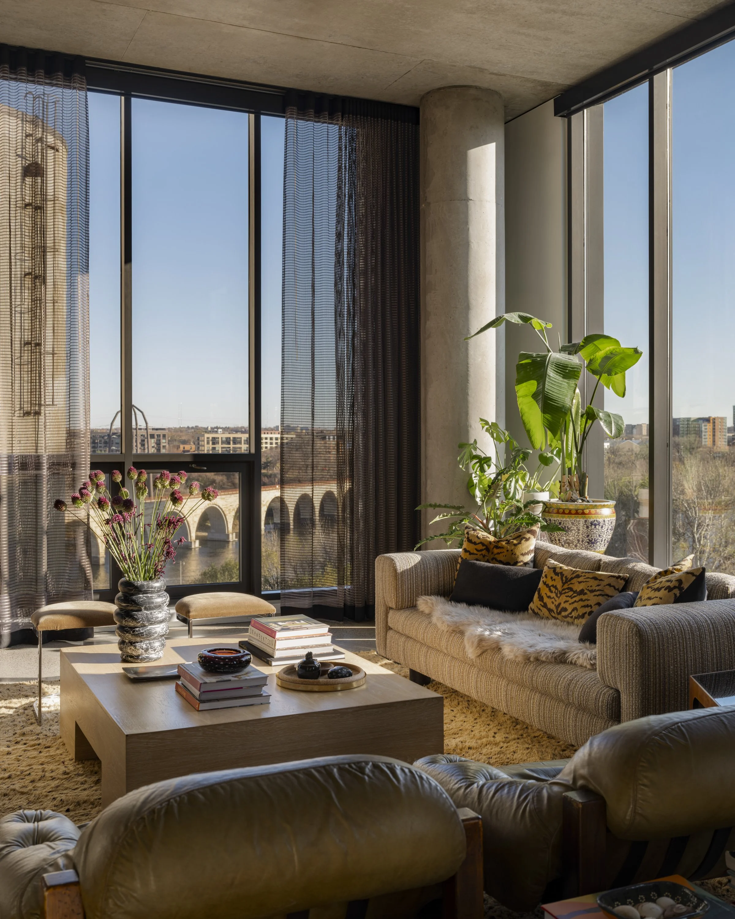 Sunlit living room with large windows, plants, a striped sofa with patterned pillows, a wooden coffee table with books, vases, and decorative items, and two lounge chairs with curved backs.