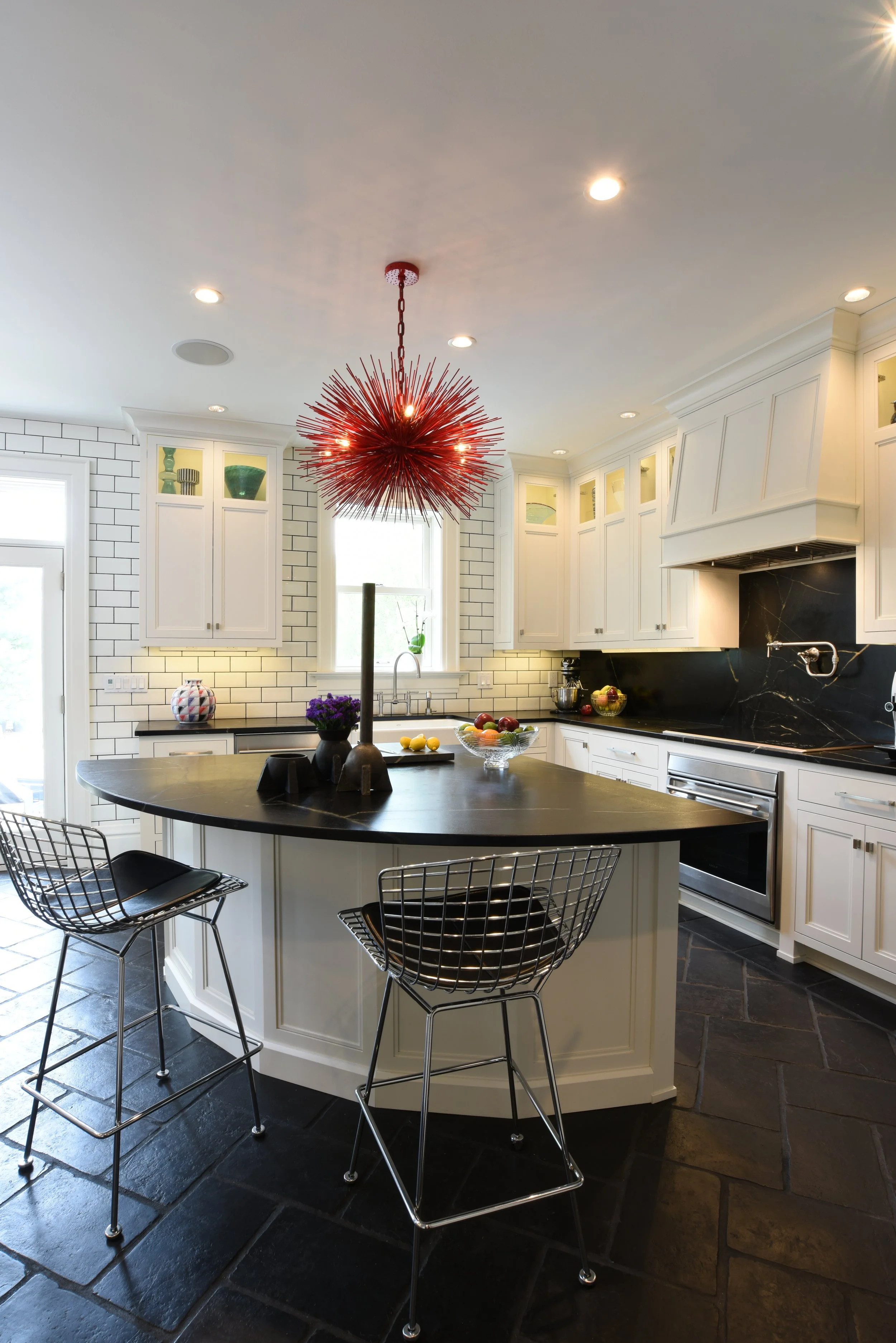 Modern kitchen with white cabinets, black countertops, a black island with three wire chairs, a red spiky pendant light, white subway tile backsplash, black slate flooring, and decorative items including a fruit bowl and vases.