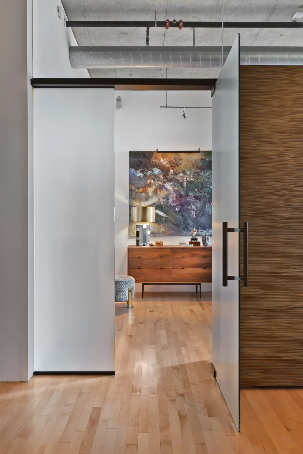 View of a modern interior with a wooden sideboard, large abstract painting, table lamp, and decorative items, seen through a partially open glass sliding door in an industrial-style space with exposed ductwork.