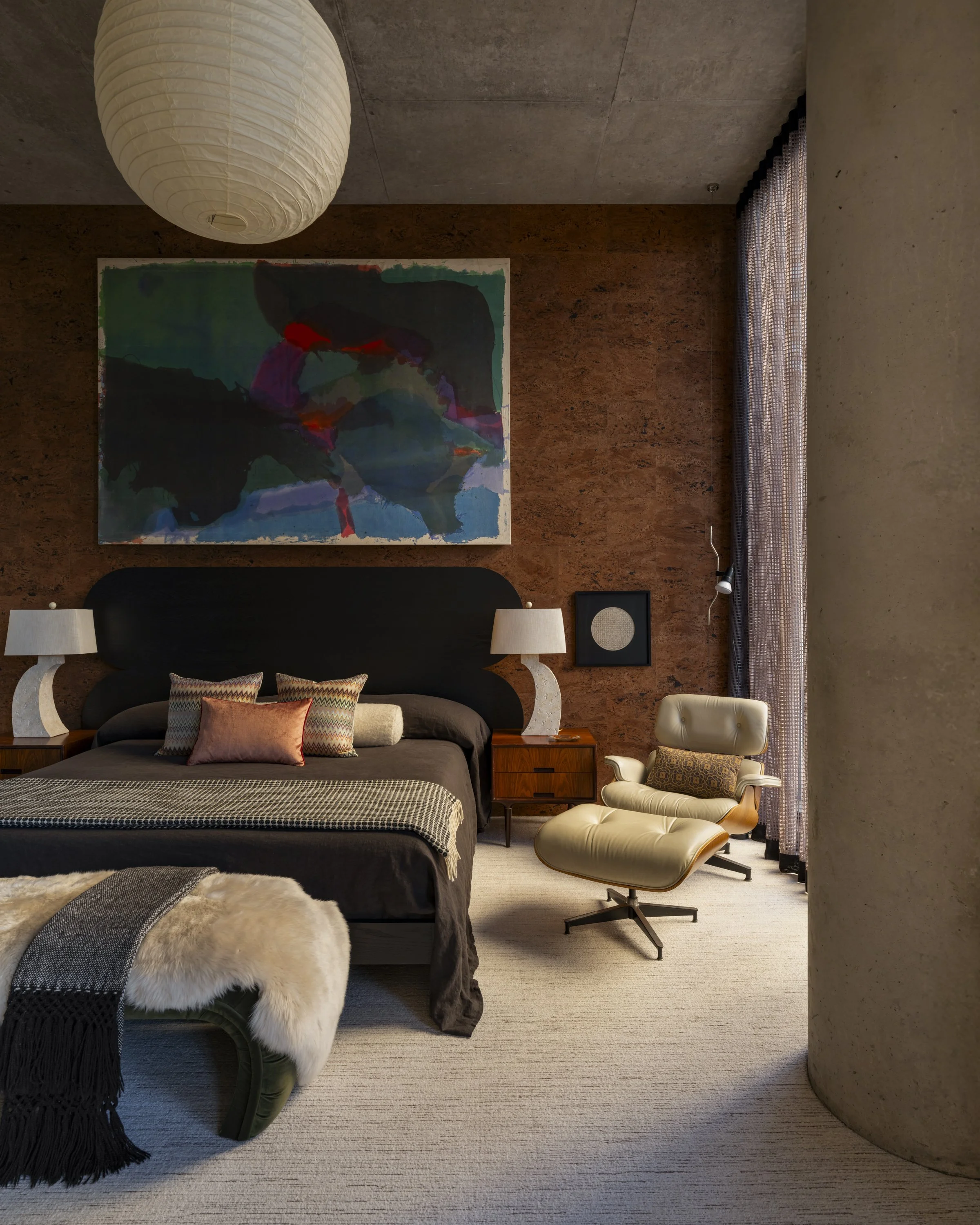 Modern bedroom with dark bed, colorful abstract artwork on brown textured wall, white hanging paper lantern, cream armchair, side tables with white lamps, window with sheer curtains, and beige carpeted floor.