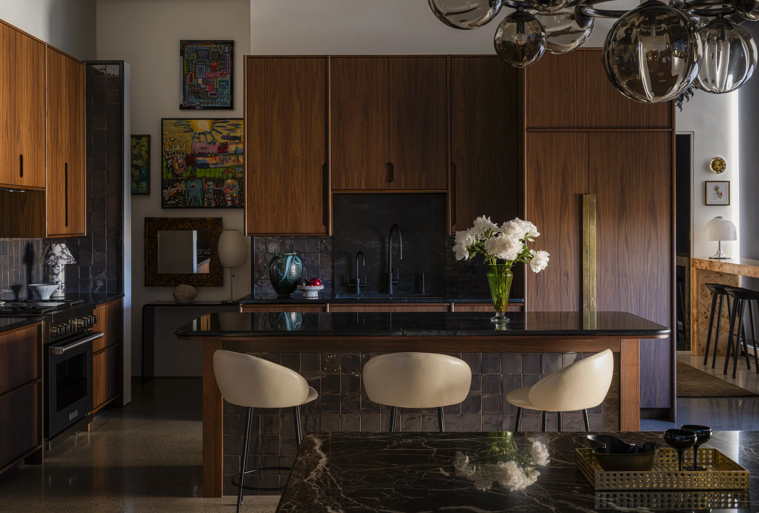 A modern kitchen with wooden cabinetry, black countertops, a black backsplash, and a kitchen island with a vase of white flowers. There are three cream-colored bar stools at the island, and a hanging glass light fixture.