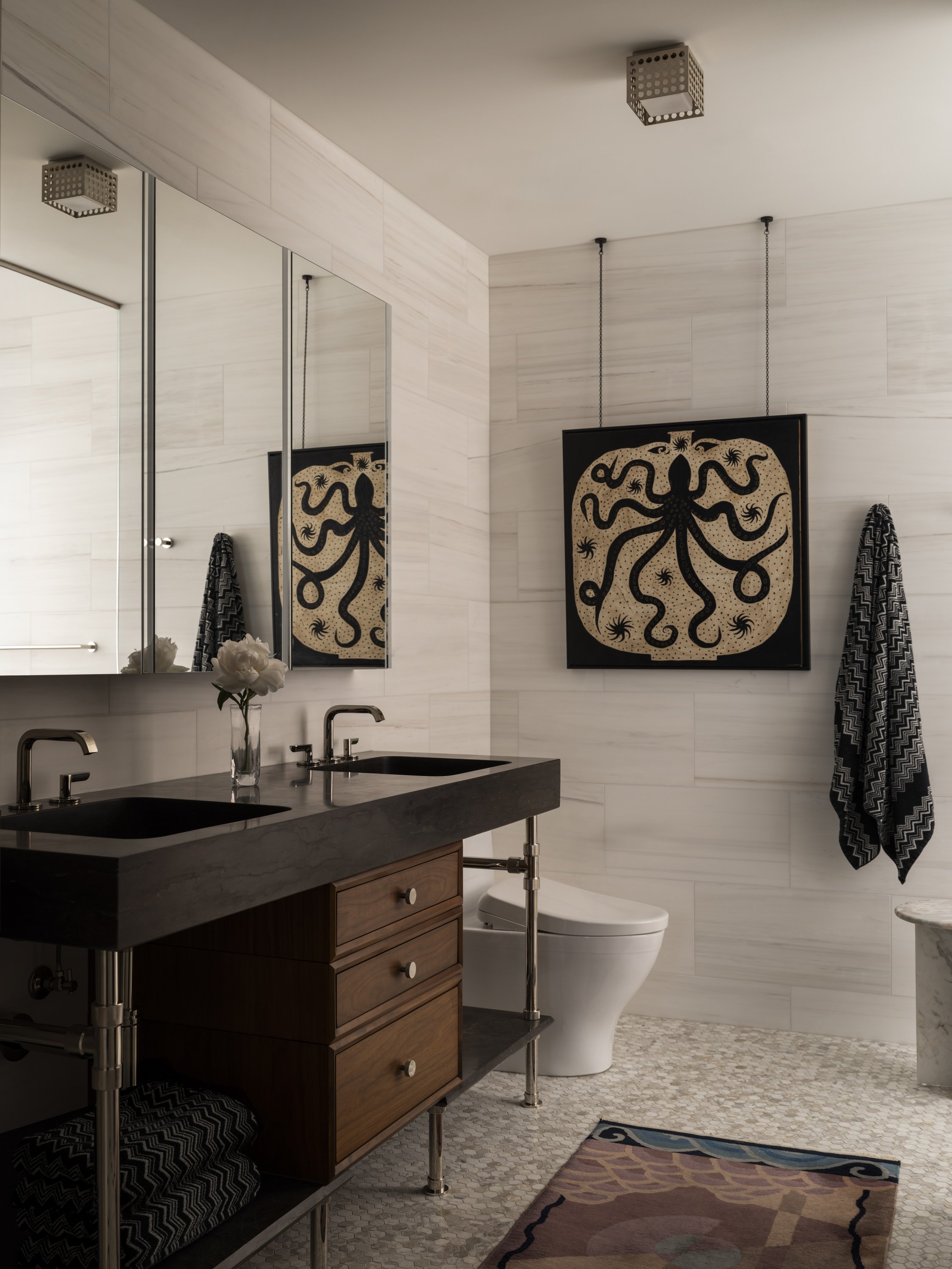 Modern bathroom with white tiled walls, dual sinks with black countertops, wooden cabinet, large mirror, black framed artwork of an octopus, towel hanging on the wall, and textured floor and rug.