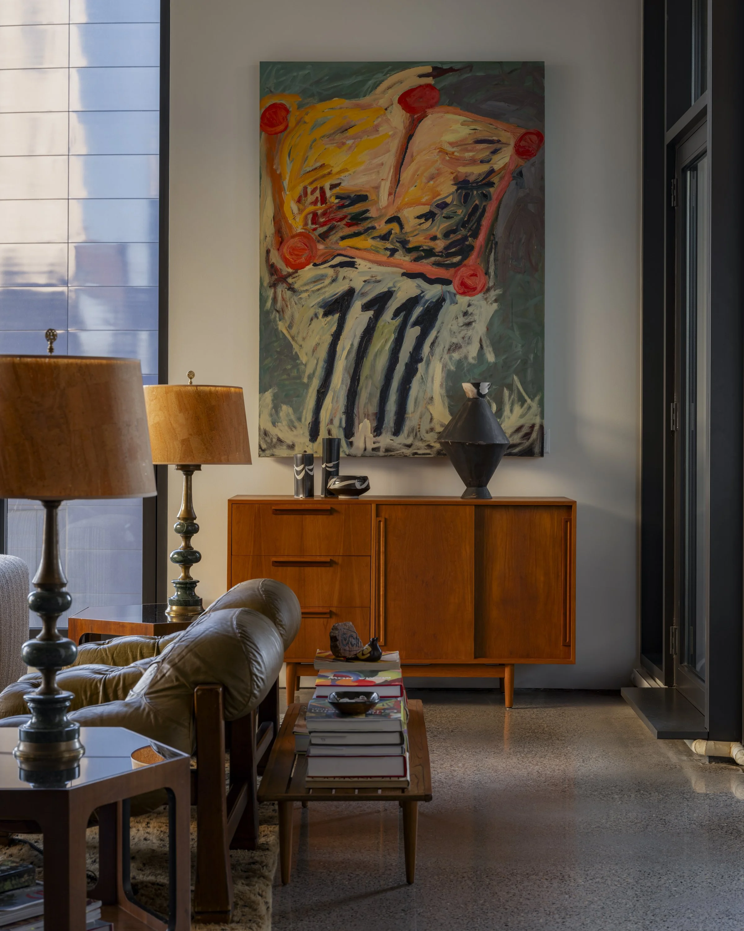 Interior of a living room with a wooden sideboard, a colorful abstract painting on the wall, table lamps, and a stack of magazines on a small table.