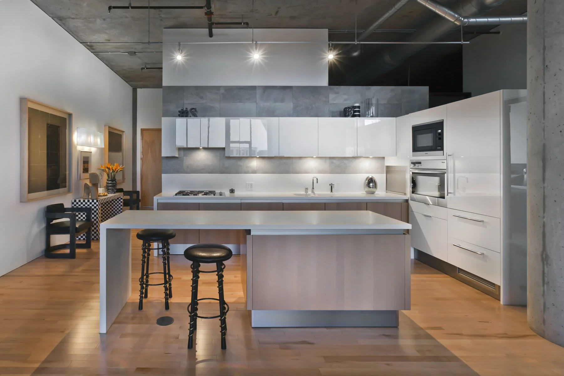 Modern kitchen with white cabinets, a central island with black stools, stainless steel appliances, wood flooring, and decorative items on the wall and counter.