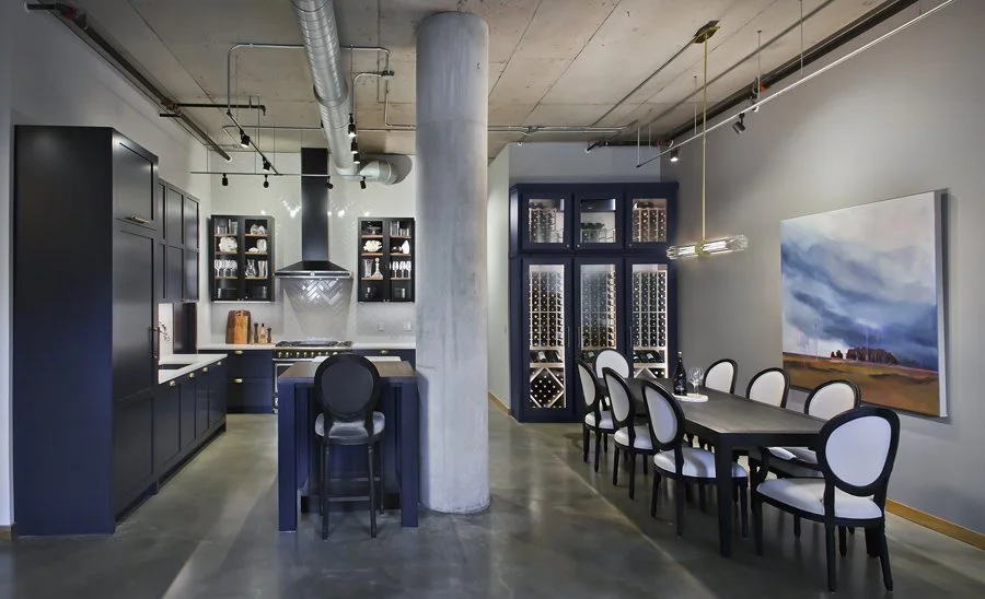 Modern kitchen and dining area with black cabinetry, a central island with barstools, and a dining table with white chairs. There are built-in wine racks and a large abstract painting on the wall.
