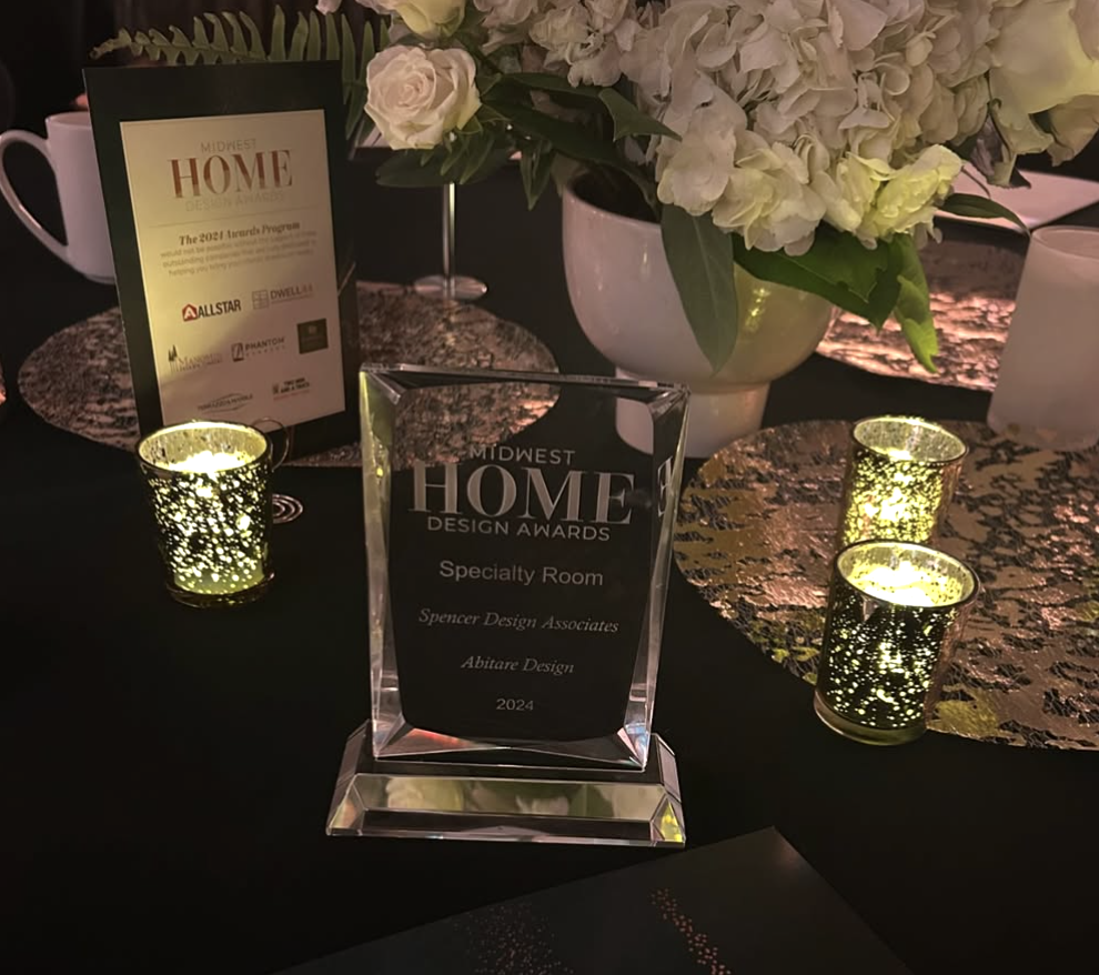 A floral centerpiece with white roses and hydrangeas, candle holders with lit candles, and award plaques on a black tablecloth at the Midwest Home Design Awards with a First Place Award to Spencer Design Associates.