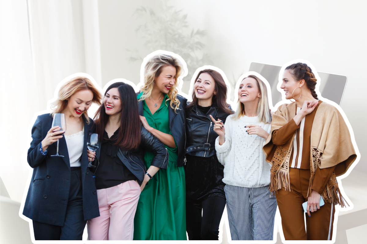 Six women standing together, smiling, laughing, and enjoying drinks at a social gathering indoors.