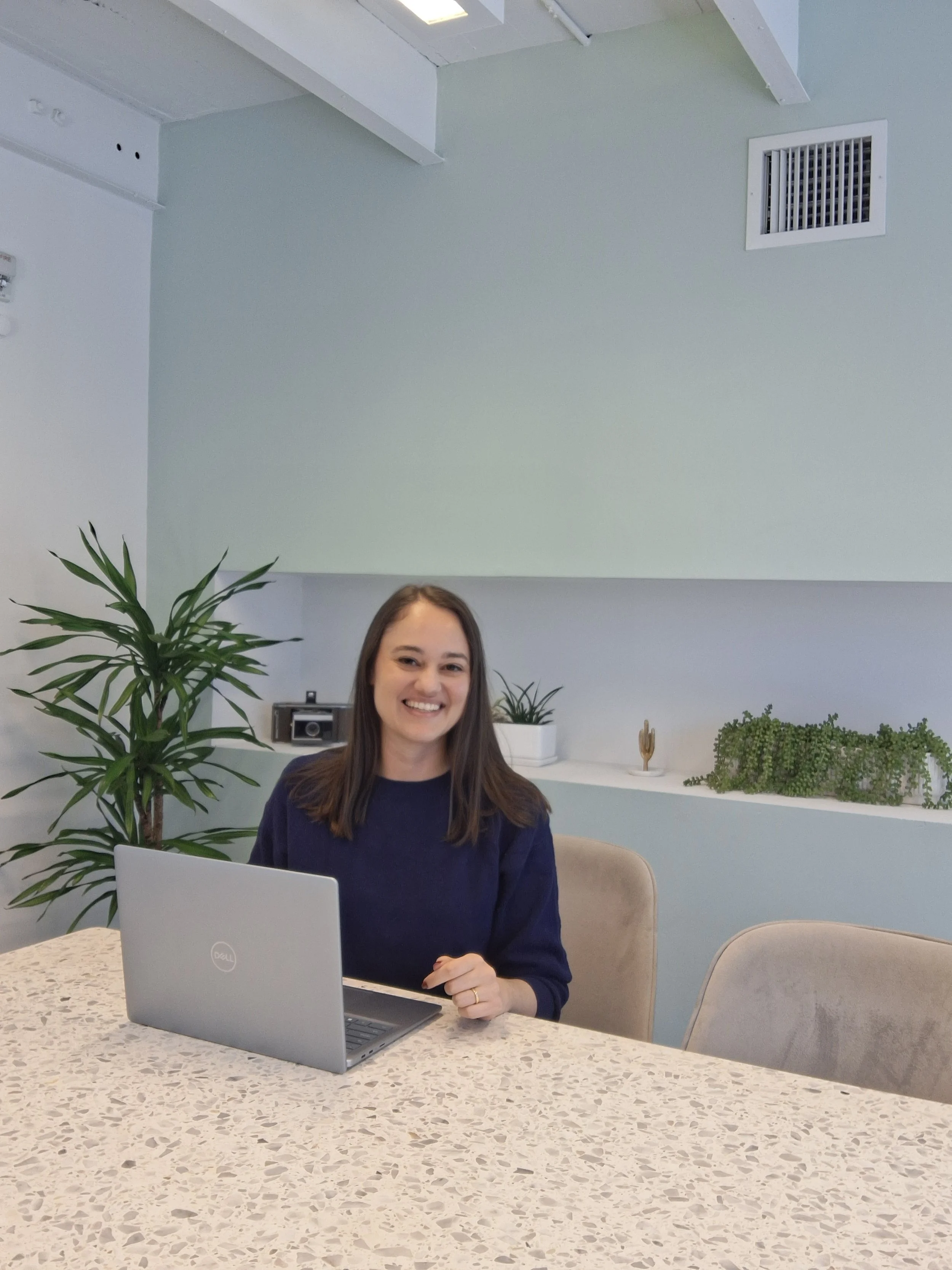 A woman with long brown hair and a navy blue sweater sitting at a desk in a modern office space with a laptop, plants, and decorative items in the background.