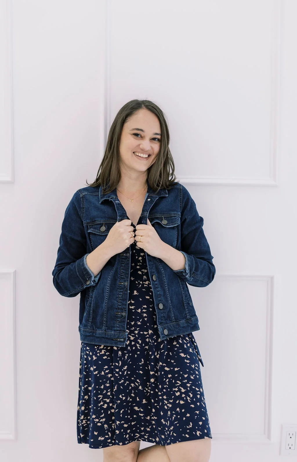 A woman smiling and holding a denim jacket in front of a plain white wall, wearing a patterned navy dress.