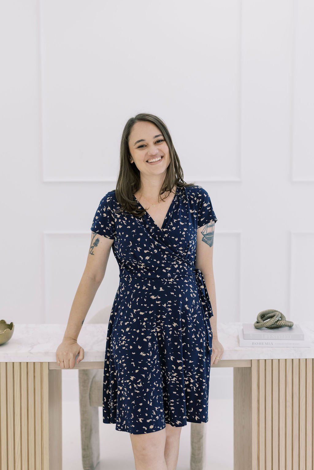 A woman with shoulder-length brown hair, wearing a navy blue dress with a white pattern, smiling and standing in a white room with modern decor on a wooden table.