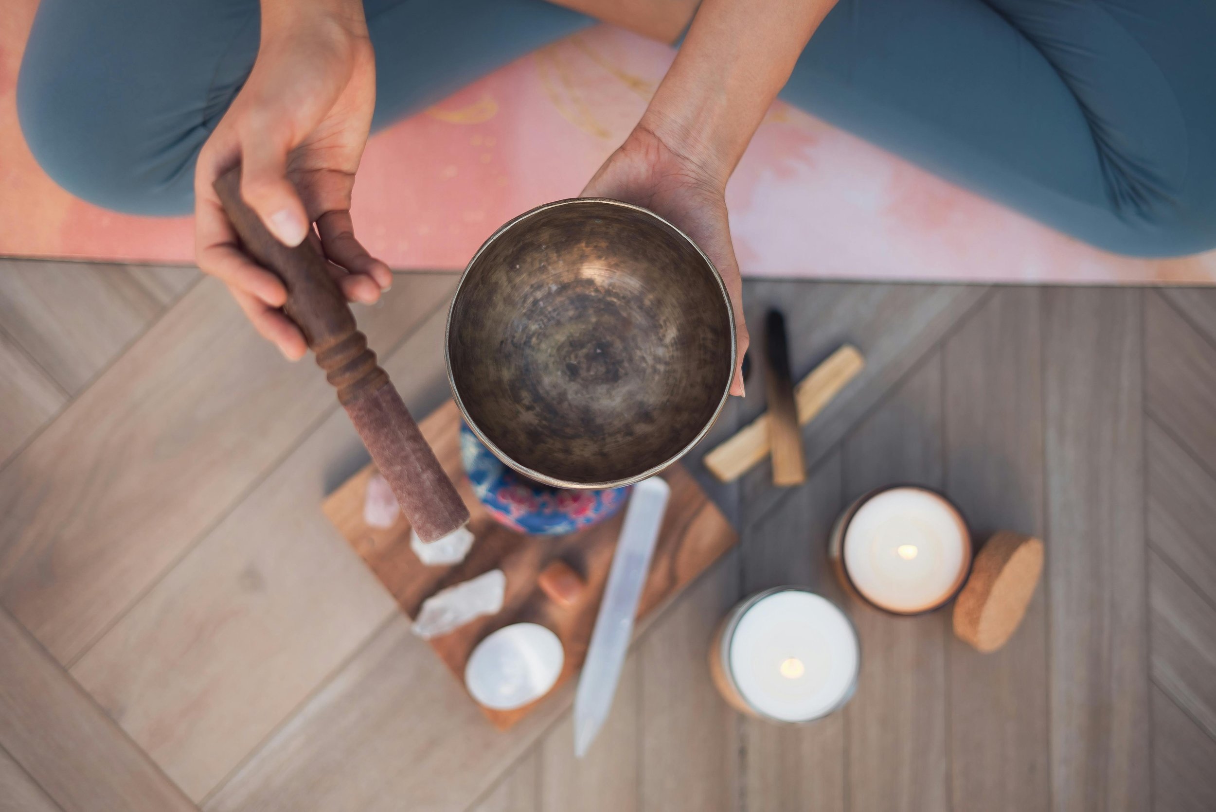 Top-down view of a person holding a singing bowl with a wooden mallet, surrounded by candles and bricks on a wooden surface.