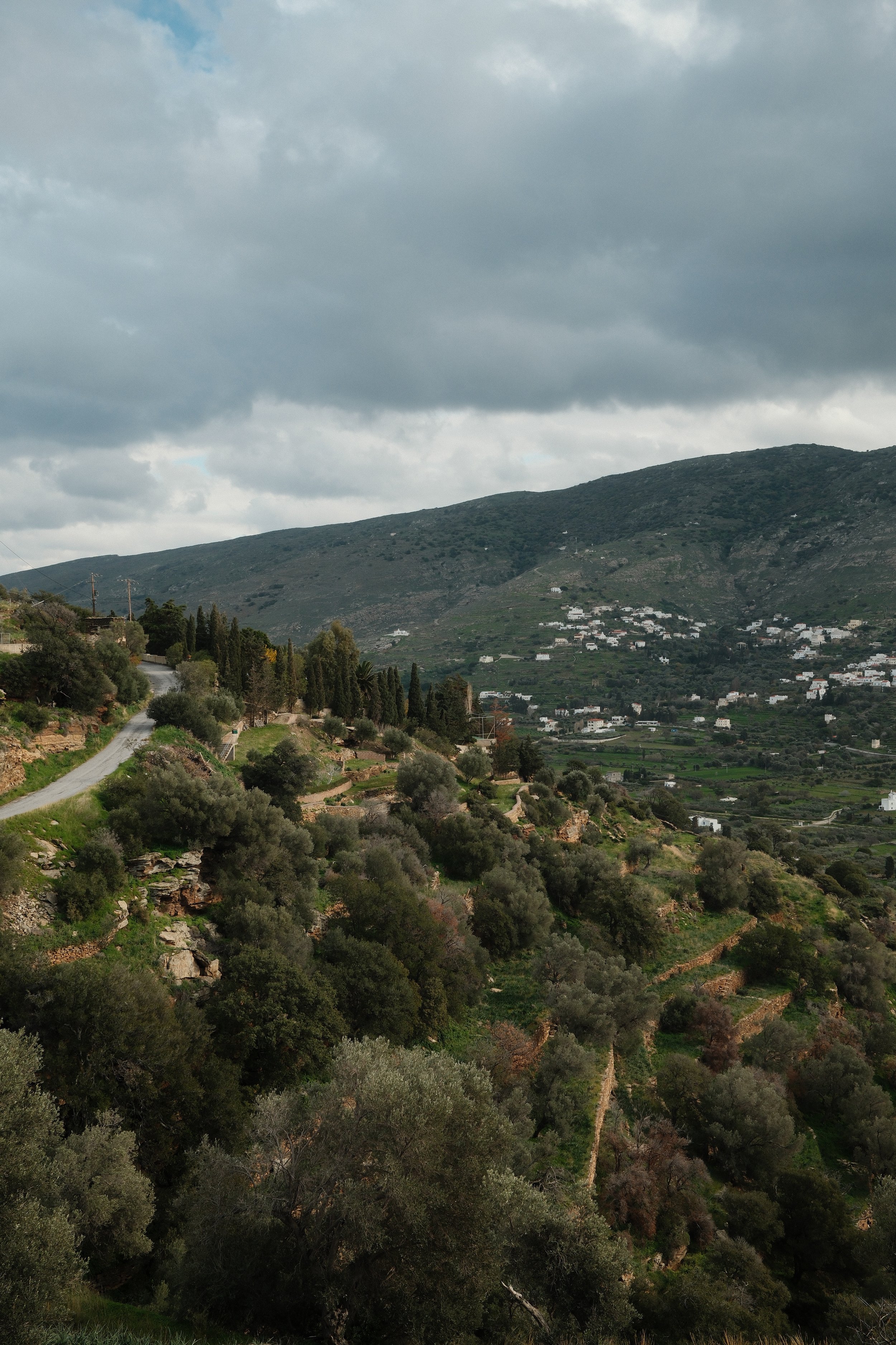 Udsigt over bjergrigt landskab med veje, træer og spredte hvide huse under en mørk grå sky.