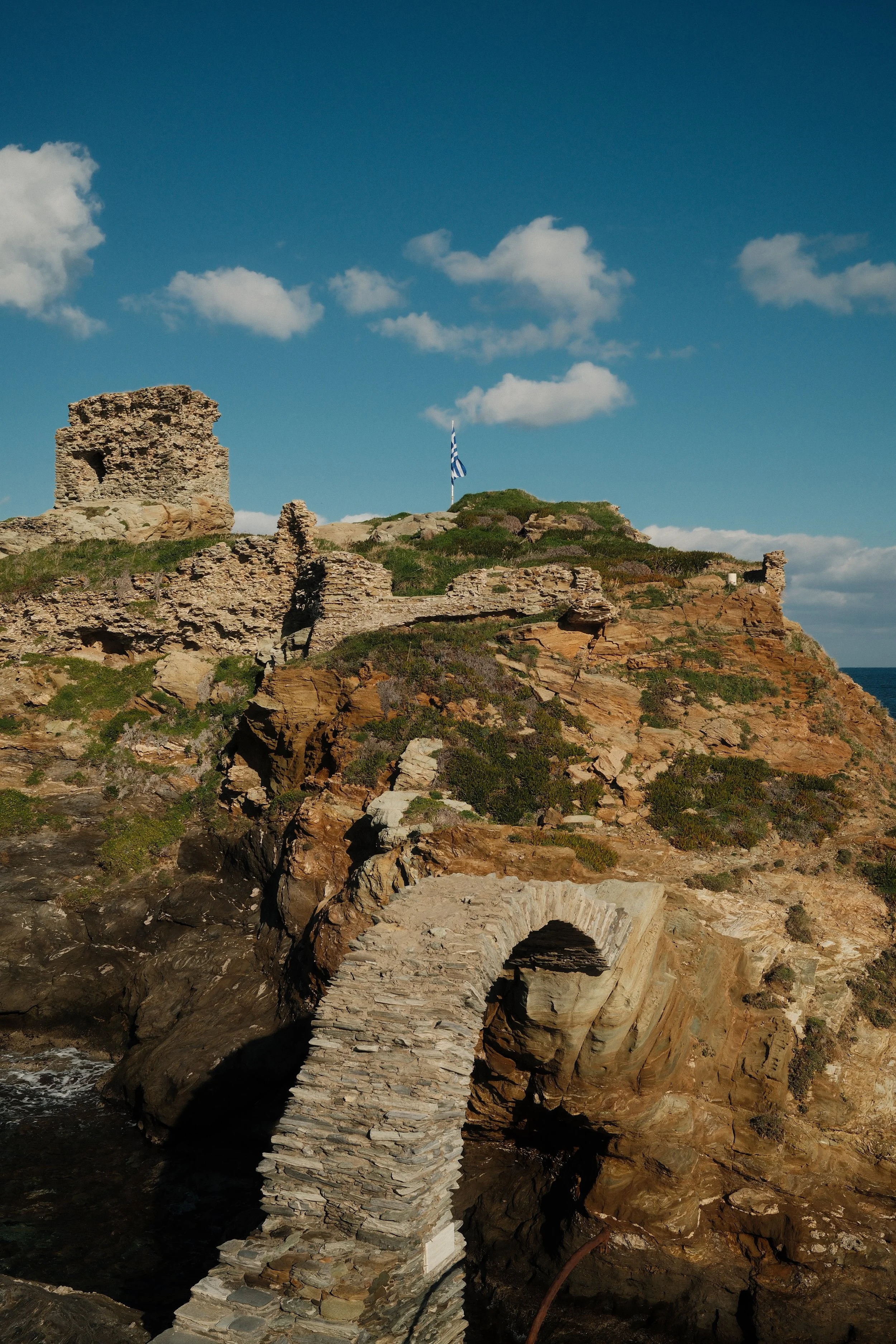 Ruiner og en bro på en klippe, med en græsk flagstang på toppen, under en blå himmel med spredte skyer.