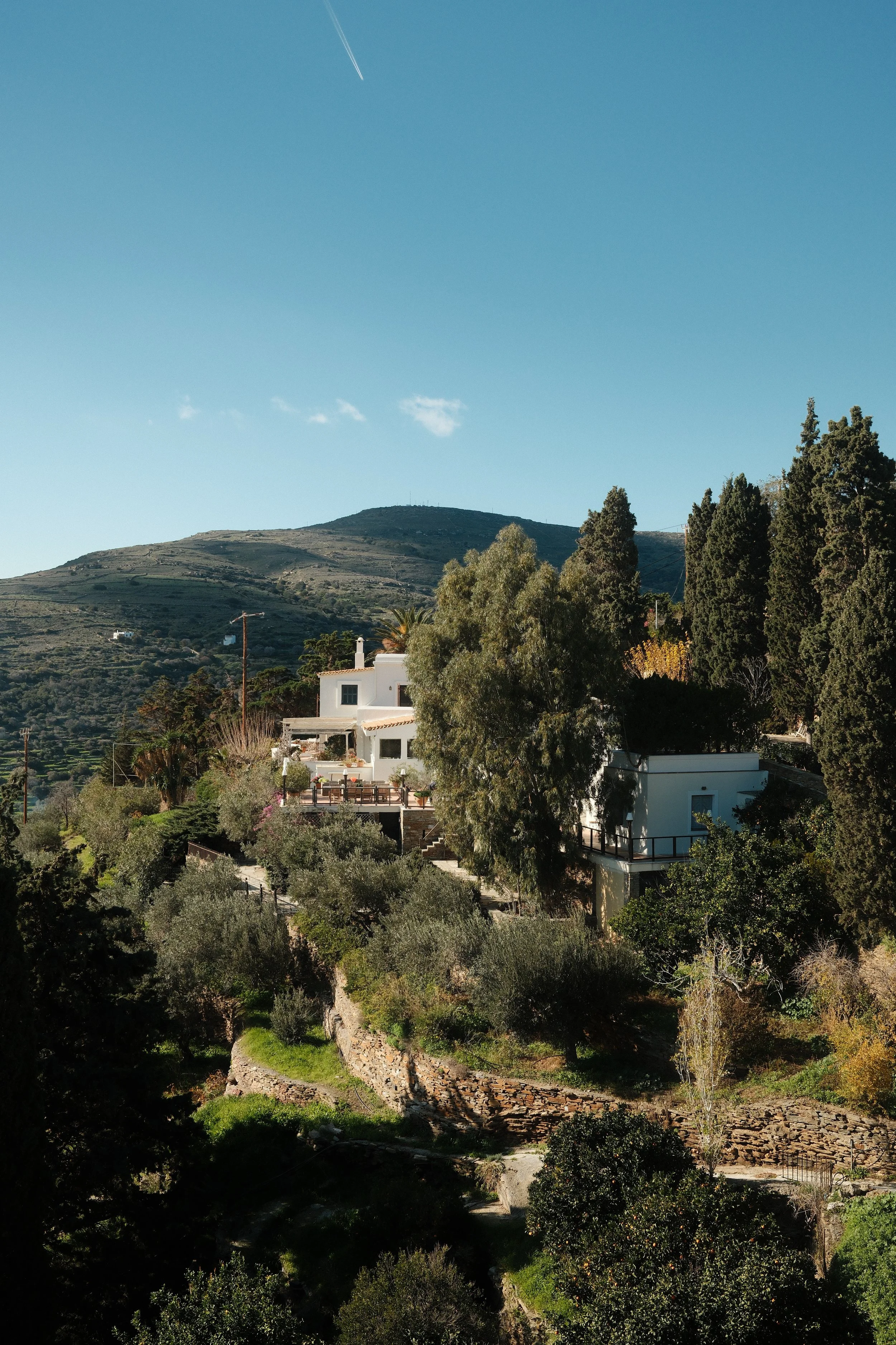 Billedet viser en rolig landsby med hvide huse omgivet af grønne træer og en bakketop i baggrunden under en klar blå himmel.