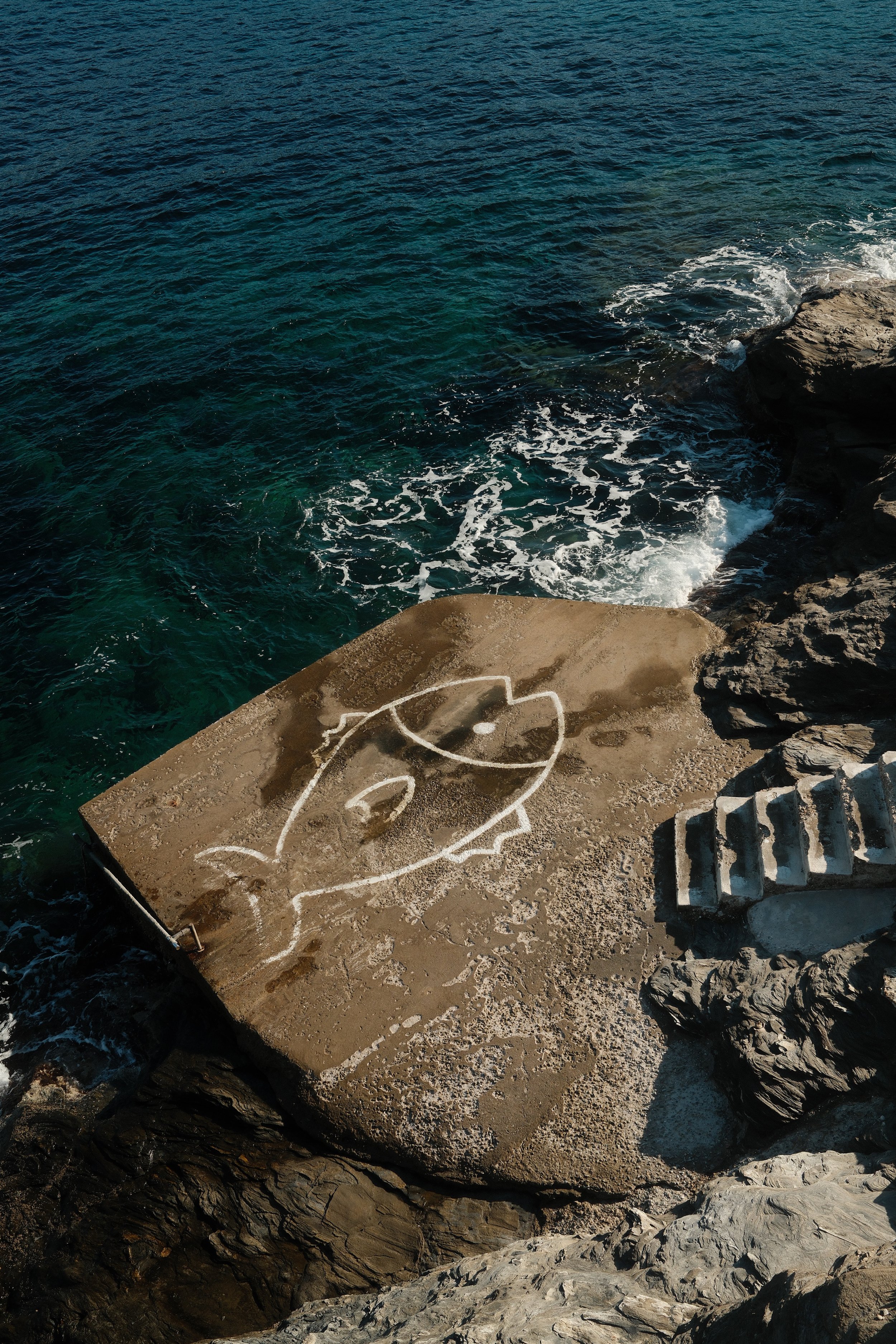 Graffiti af et ansigt på en betonplatform ved havet med klipper omkring, set fra oven.
