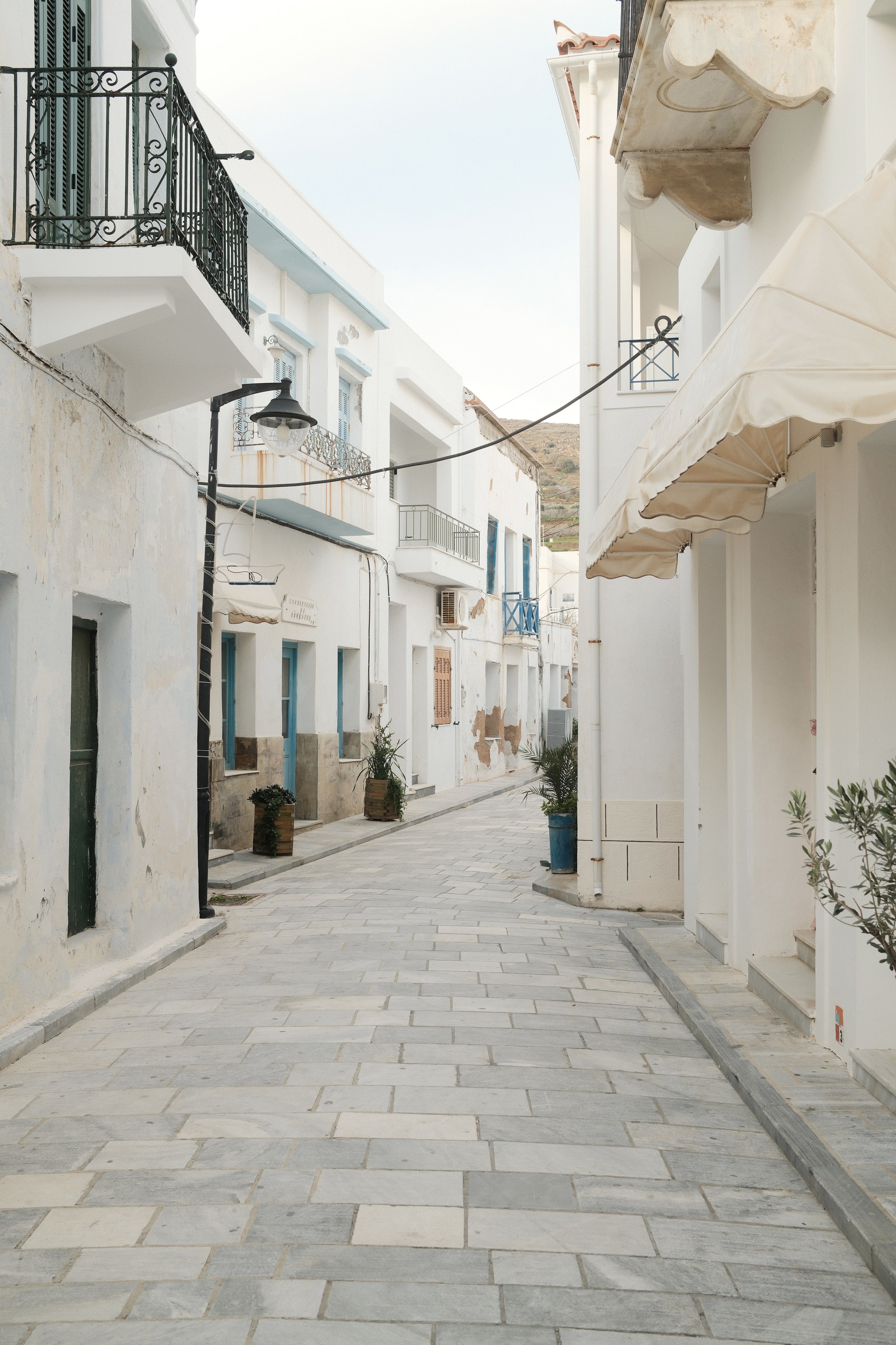 En tom, snoet gade i en by med hvide bygninger, nogle med balkoner og vinduer med blå rammer, og små planter i potter langs fortovet. Der er en gadelampe og nogle solskærme, og bjerg i baggrunden.
