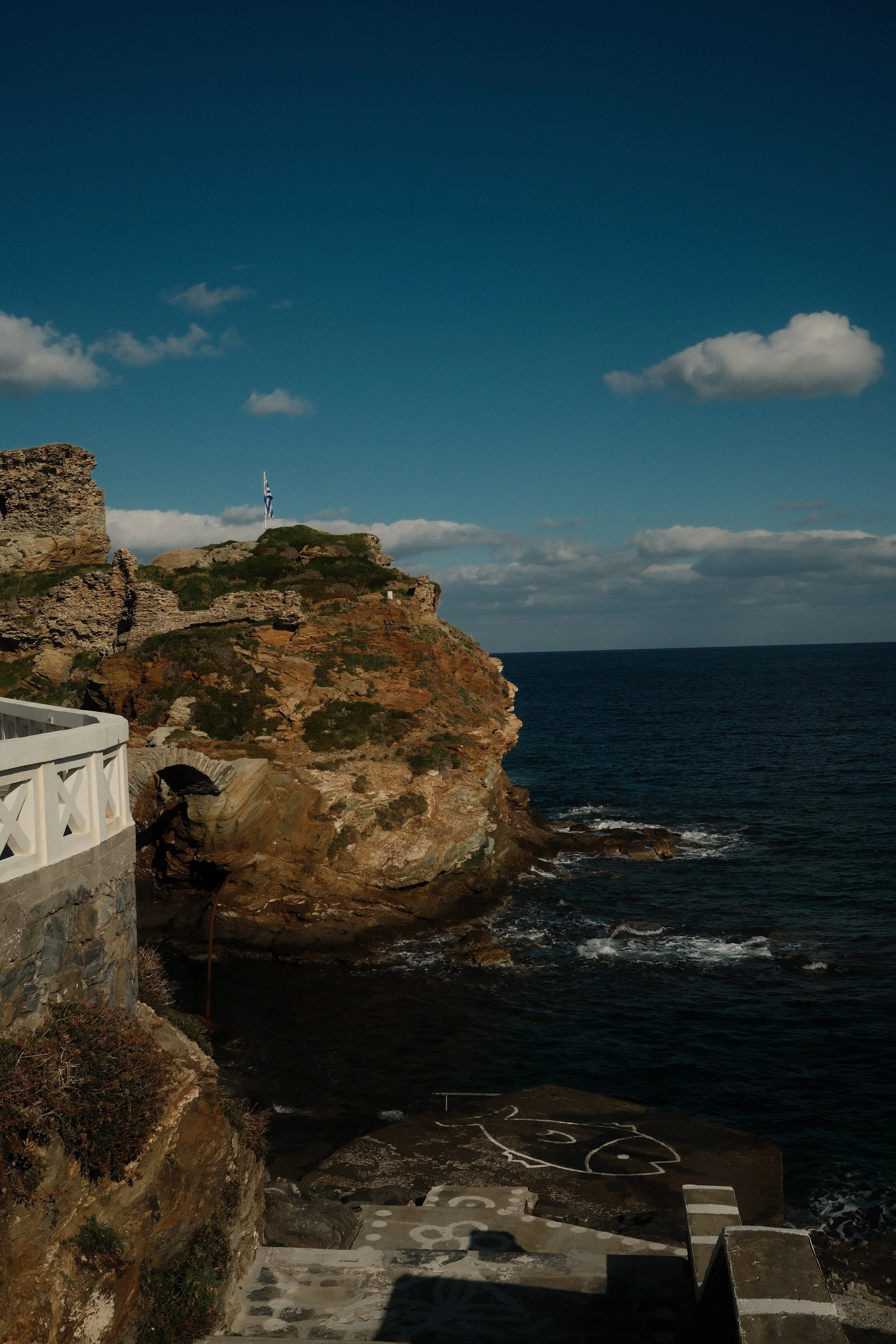 Udsigt over en klippe ved havet med en græsk flag på toppen og en hvid trappe i forgrunden.