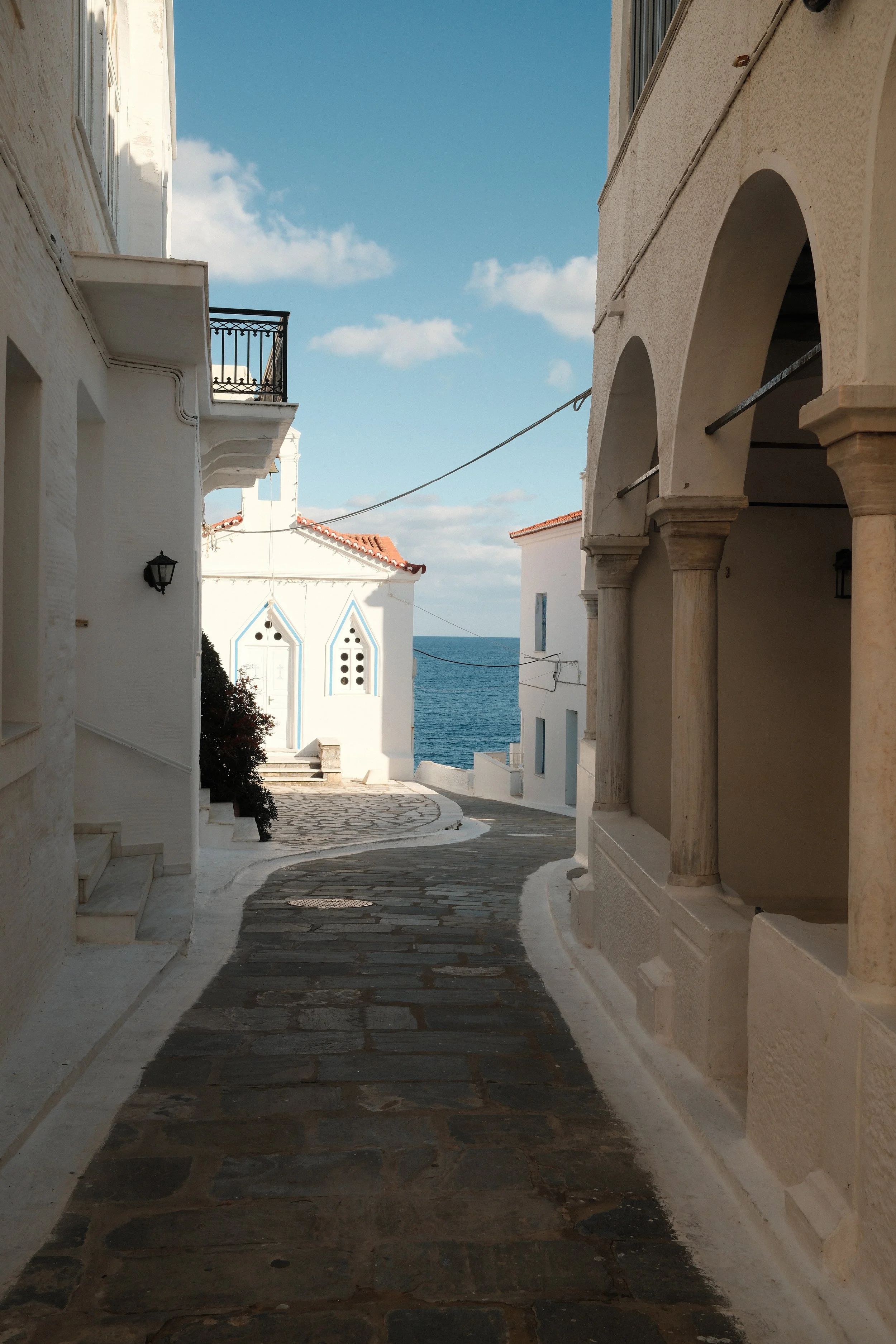 En smal, brolagt vej i en hvidkalket landsby med bjergtagende udsigt over havet, omgivet af hvide bygninger med buede arkader og balkoner.