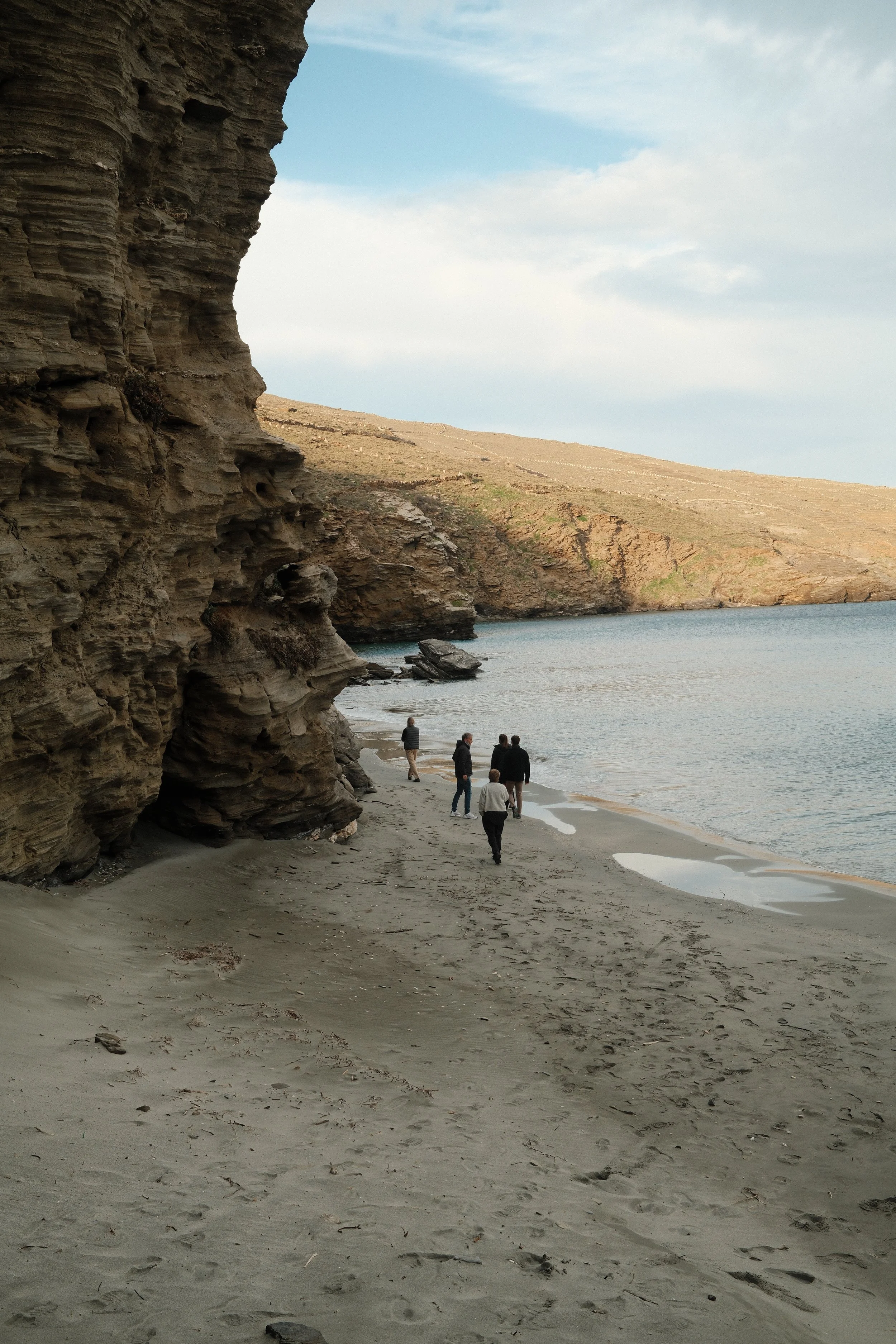 En gruppe mennesker går langs en kystlinje med klipper og sandstrand, under en overskyet himmel.