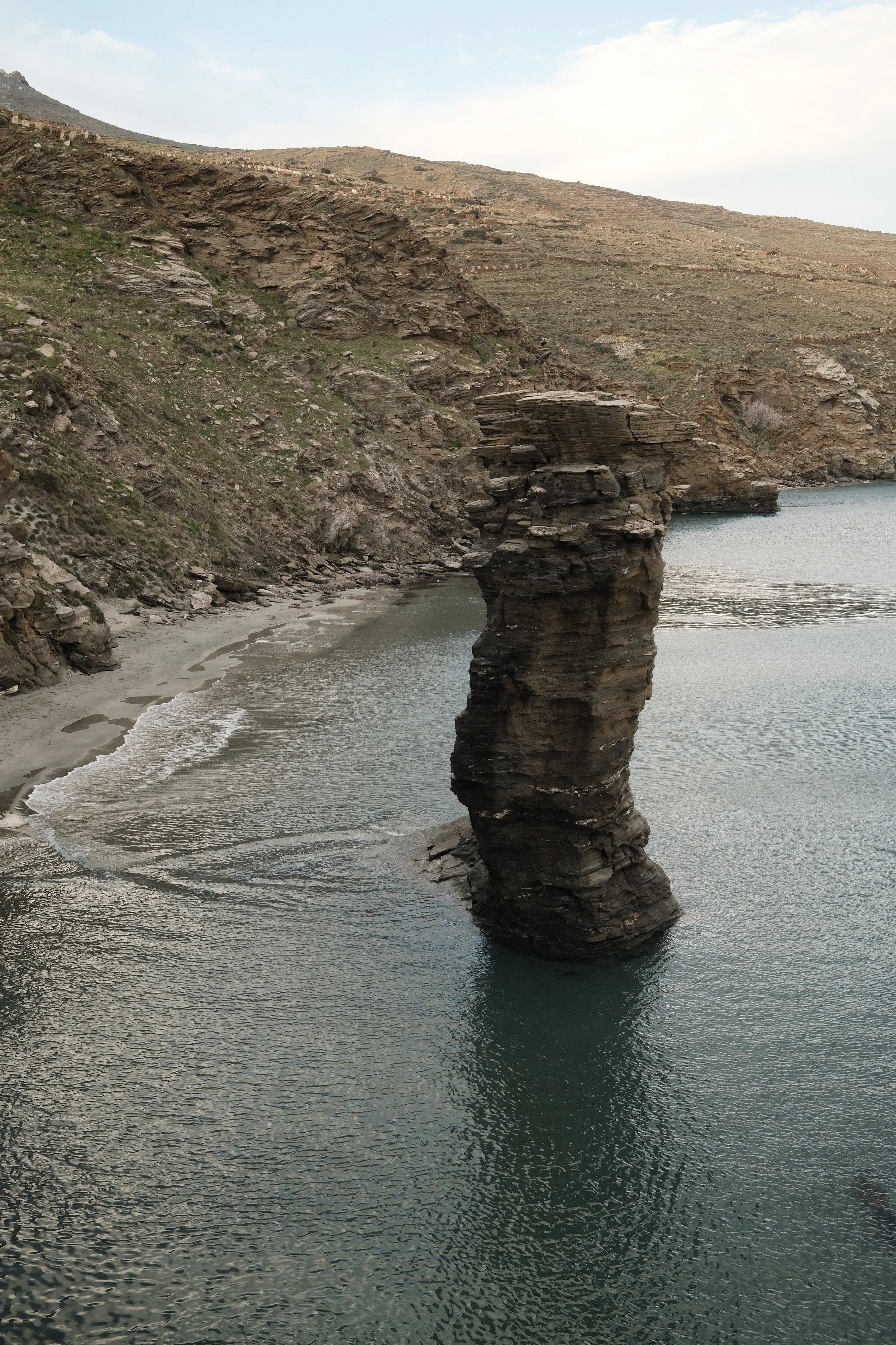 En stor, lodret klippeformation i vandoverfladen ved en kystlinje med klippebakke i baggrunden.