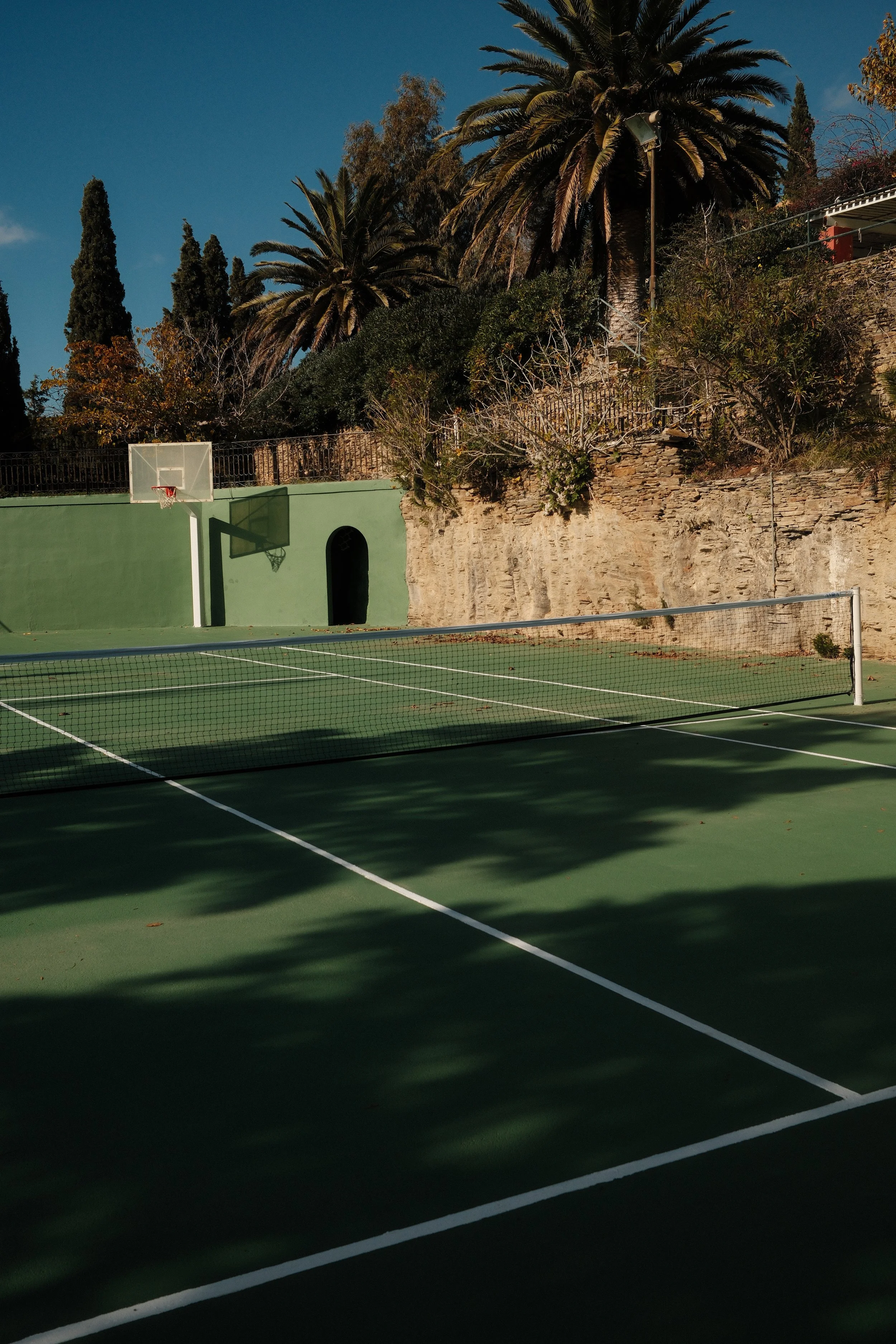 En udendørs tennisbane med grøn overflade og hvide linjer, indhegnet med net, under skygger fra træer, med en basketballkurv og klippevæg med buske og palmer i baggrunden.