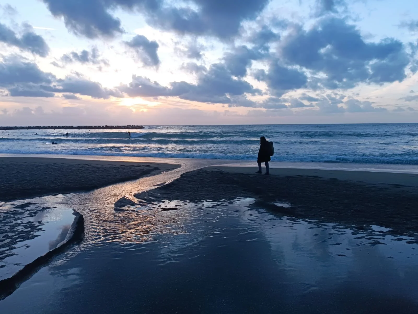 Persona che guarda il mare sulla spiaggia al tramonto o all'alba, con nuvole nel cielo e onde che si infrangono sulla riva.