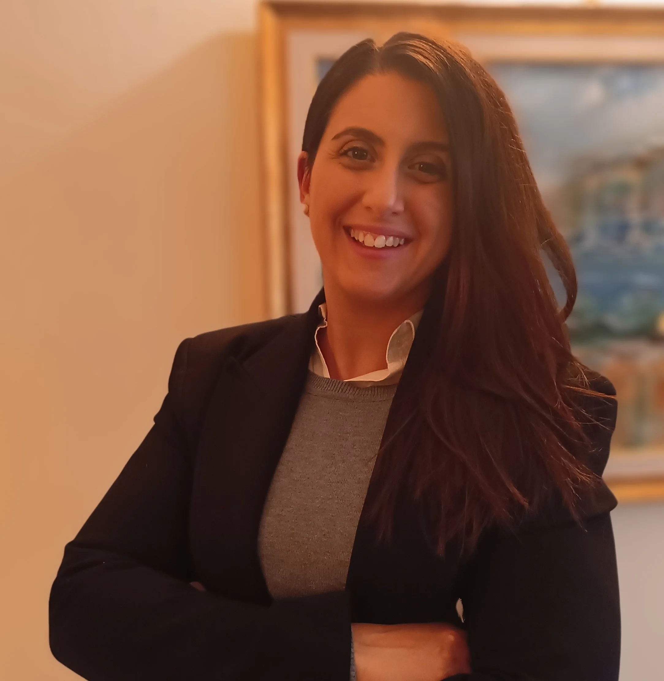 Una donna sorridente con capelli castani, vestita in abiti formali, in un ambiente interno con dipinti sullo sfondo.