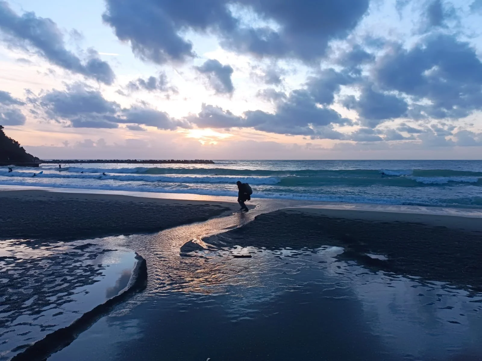 Scena di un tramonto sulla spiaggia con nuvole sparse nel cielo, onde che si infrangono e una persona che si china vicino all'acqua, mentre altre sullo sfondo camminano lungo la riva.