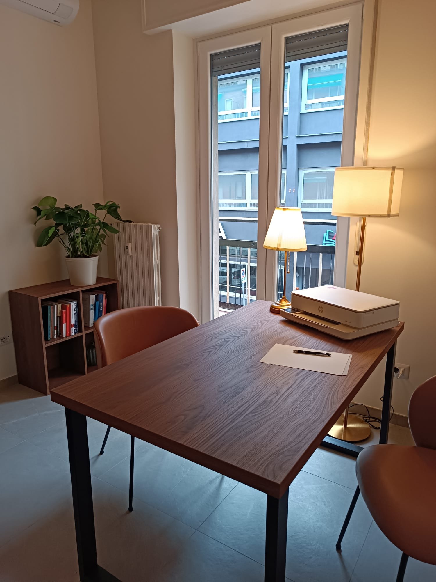 Una stanza con un tavolo in legno con una sedia di fronte, una lampada da tavolo e una lampada da terra vicina, una finestra con vista su un edificio e una pianta in un angolo.