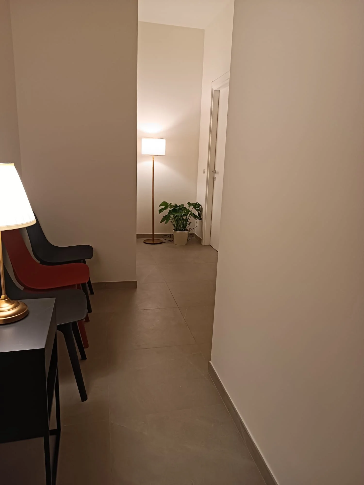 Angolo di un corridoio con pareti bianche, pavimento in piastrelle beige, lampada da terra con luce calda, pianta in vaso e sedie impilate accanto a un tavolino con lampada.