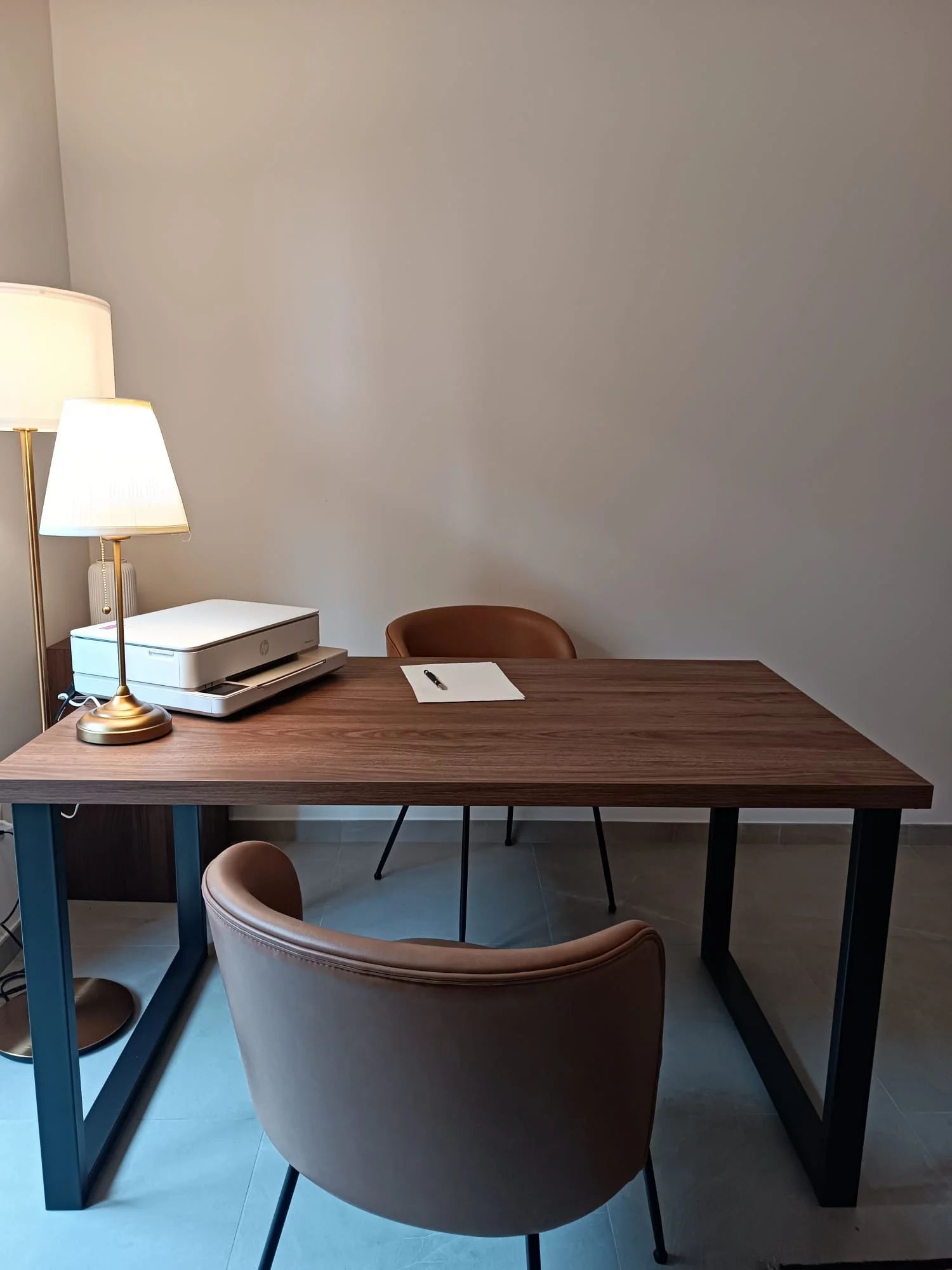 Ufficio con scrivania in legno, due sedie marroni e una lampada da tavolo. La scrivania ha una stampante e una penna su un foglio di carta.