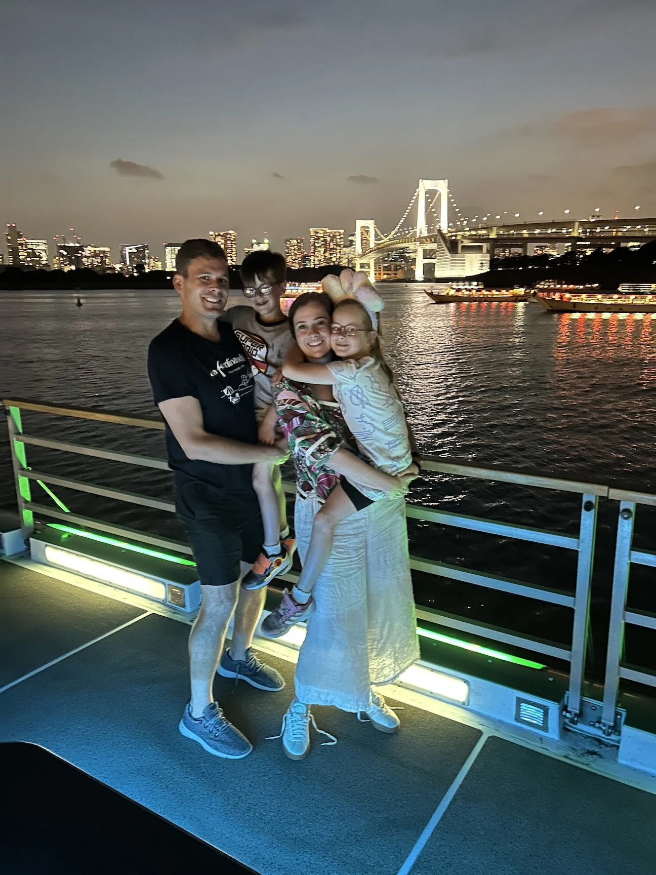 A family of four smiling and posing together on a boat with the Tokyo skyline and a lit-up bridge in the background at dusk.