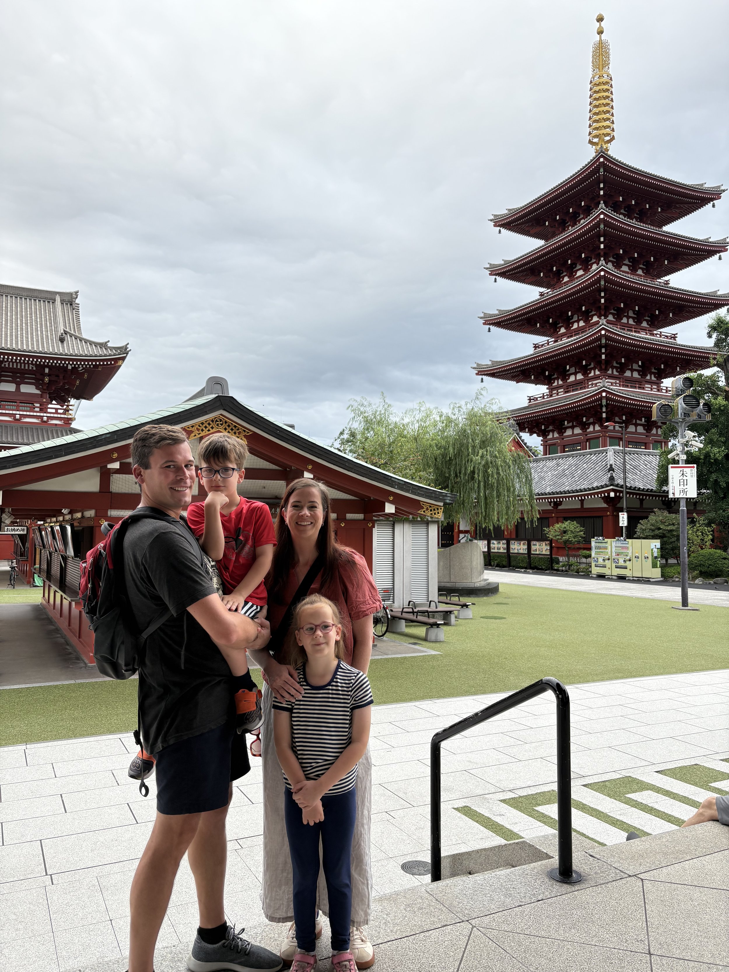 A family of four posing in front of a traditional Japanese temple, with a pagoda and temple buildings in the background, under a cloudy sky.