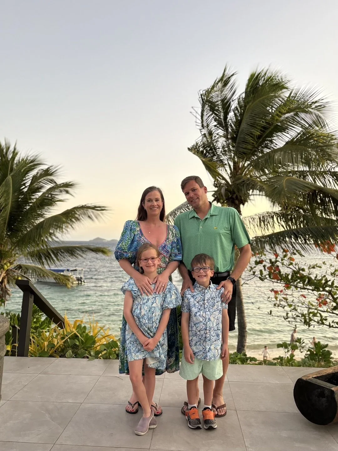 A family of four standing on a beachside patio in Fiji at sunset, with the ocean and palm trees in the background.