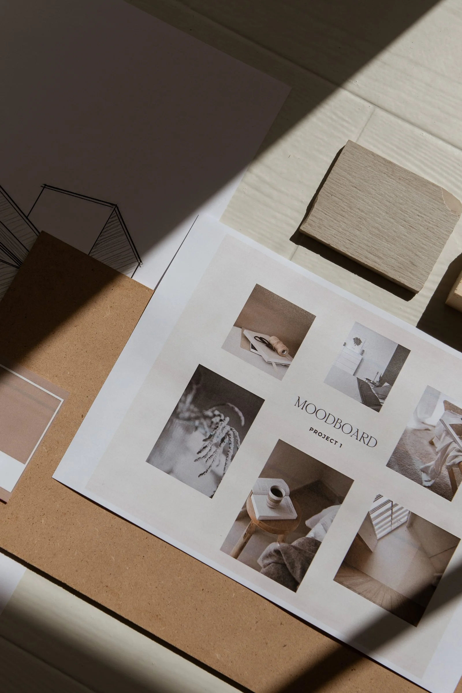 An interior design brand identity mood board titled 'Project 1' with photos of home decor and furniture, placed on a table with light wood and cork surfaces.