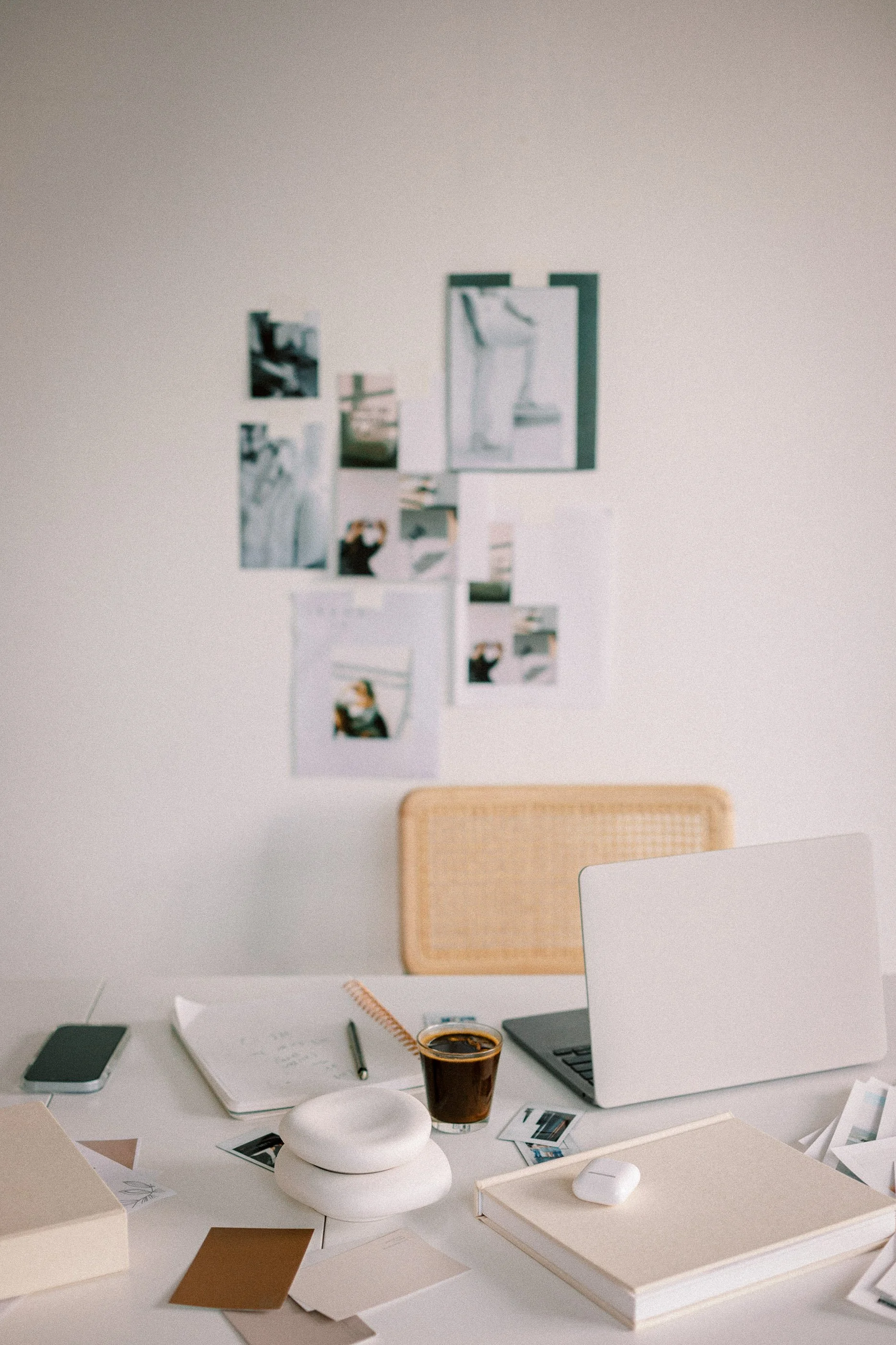 Marketing agency desk in California with open laptop, notebook, pen, coffee, mouse, photos, and books; beige chair; wall with photo collage.