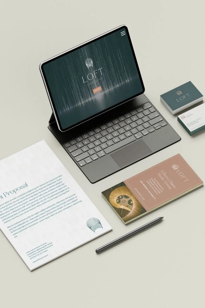 A laptop on a beige desk displaying a website with the logo 'LOFT' and a microphone graphic. Next to the laptop are business cards, a brochure titled 'LOFT', a pen, and a printed document.