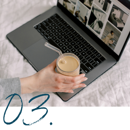 Person holding a glass of iced coffee with a straw, working on a laptop with photo thumbnails on the screen, on a white surface.