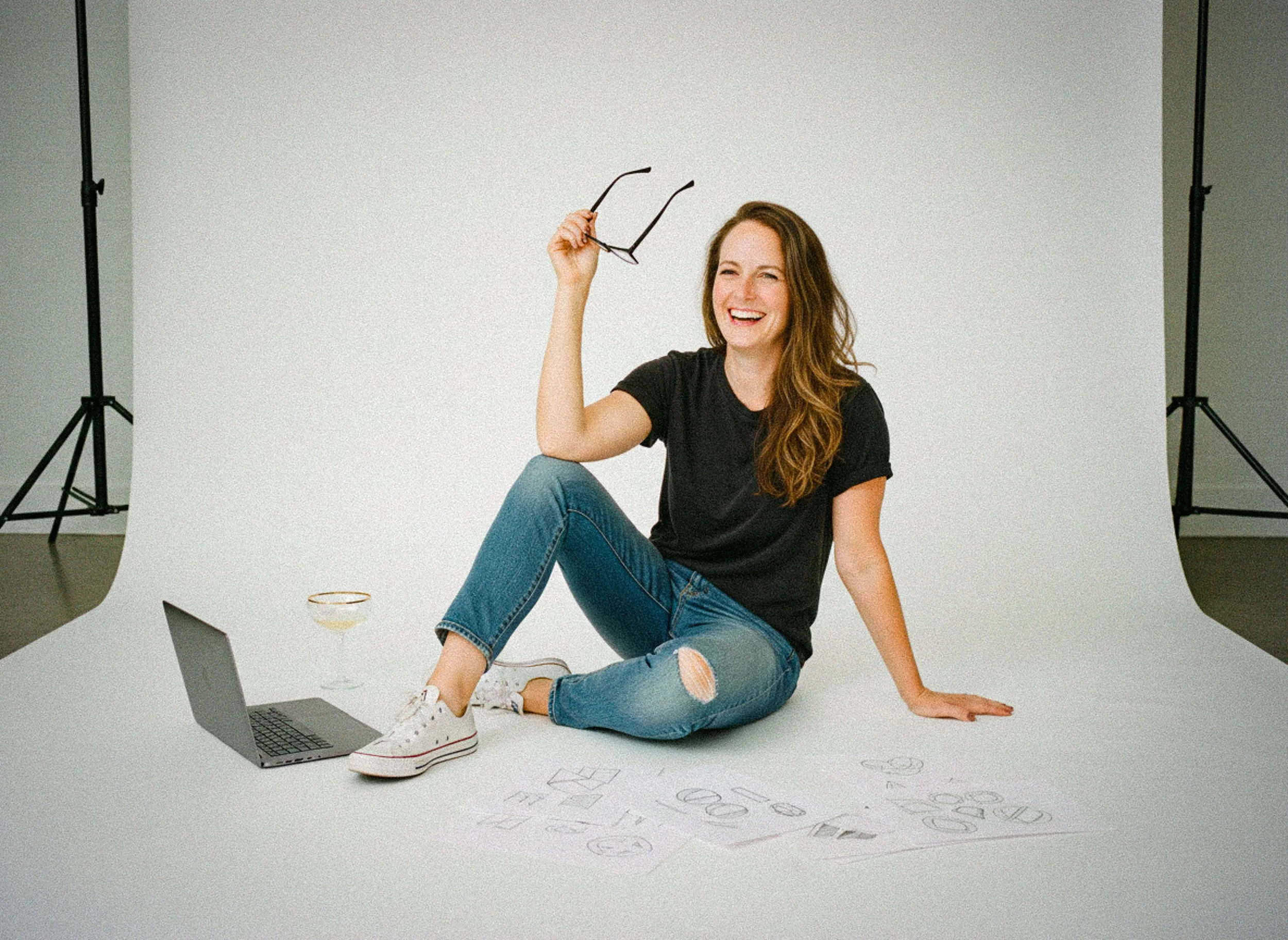 A woman with long brown hair, wearing a black t-shirt, ripped jeans, and white sneakers, is sitting on the floor in a photography studio. She is smiling and holding glasses in her right hand. There are sketches on paper, a laptop, and a glass of drink nearby.