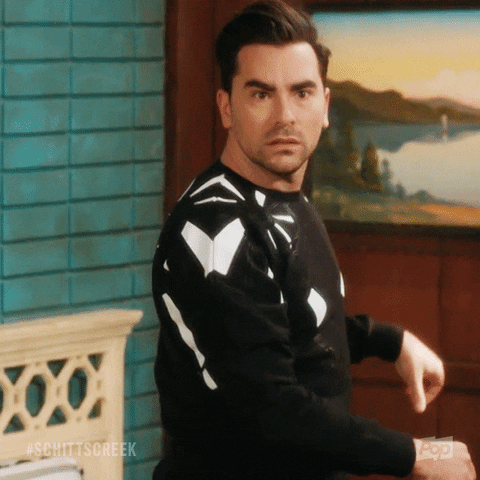 A man with dark hair and a serious expression, wearing a black sweater with white patterns, sitting in a room with teal walls and a landscape painting in the background.