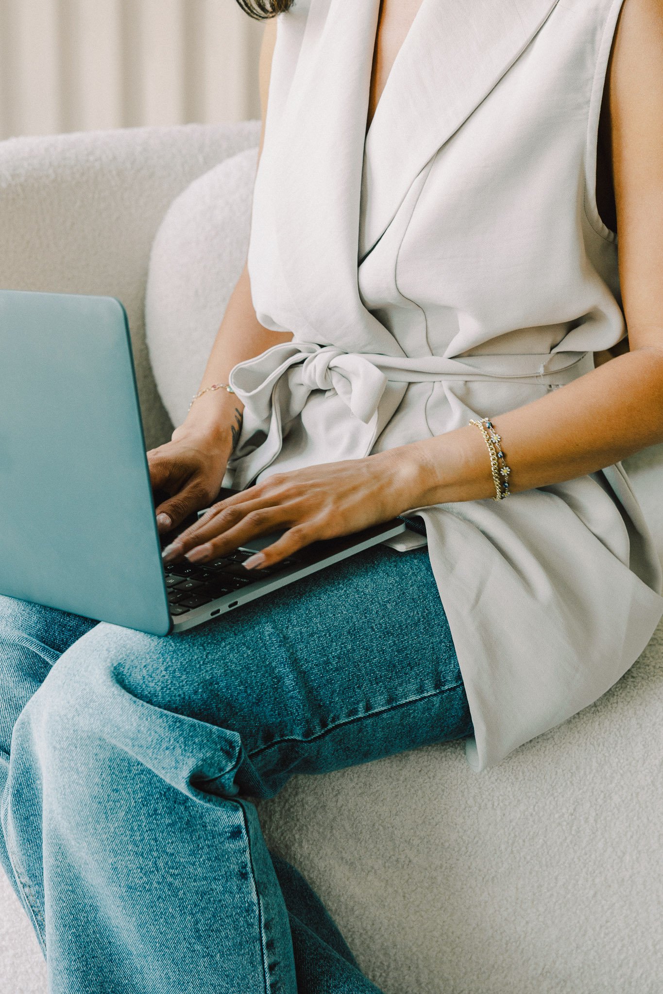Close-up of a person sitting on a beige couch, using a laptop. The person is wearing a white sleeveless top with a tie at the waist, blue jeans, and jewelry including bracelets. The focus is on their hands typing on the laptop keyboard.