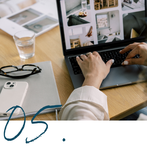 Person using a laptop to browse interior design photos, with glasses, a glass of water, a smartphone, and printed images on a wooden desk.