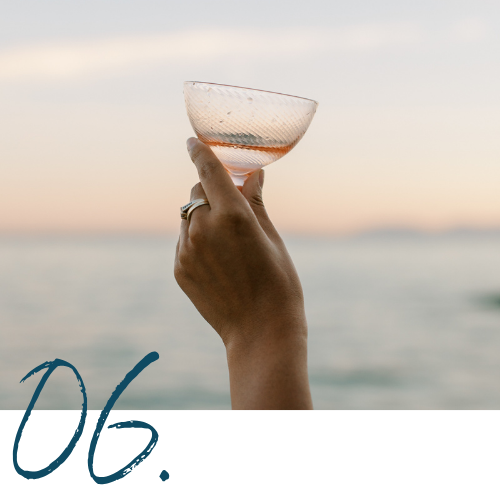 A hand holding a clear glass with a pinkish liquid, against a sunset or sunrise sky over water.