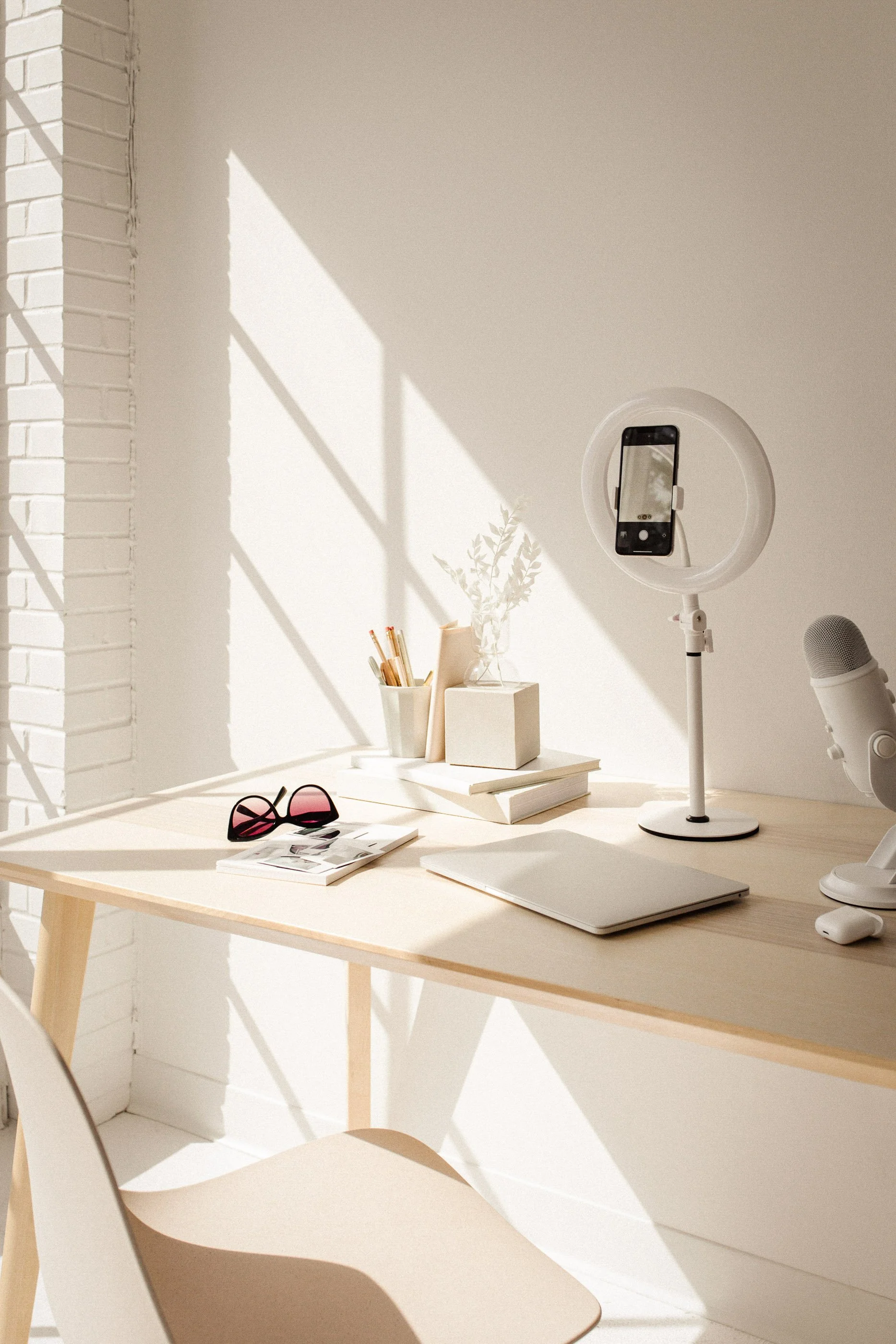 Bright workspace with a wooden desk, closed laptop, sunglasses, books, and a social media manager's ring light with a smartphone, accented by sunlight shadows.