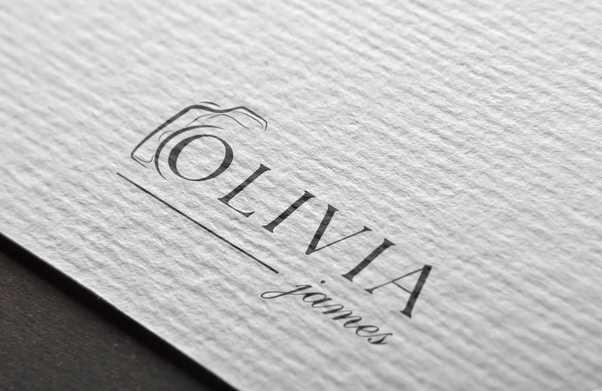 Close-up of a textured white paper with the logo 'OLIVIA james' and a camera icon above the text.