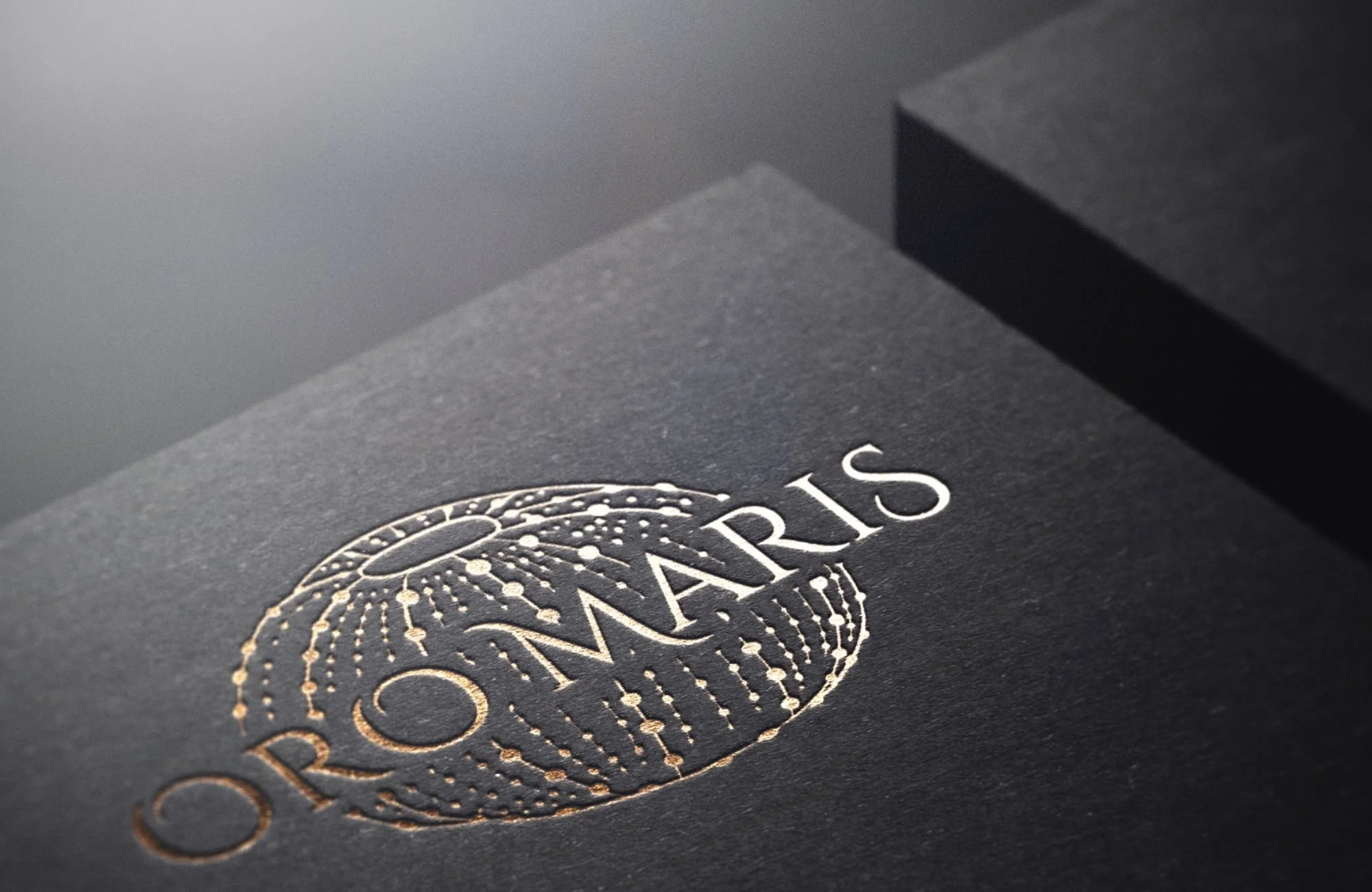 Close-up of a black textured paper with embossed gold foil text 'OROMARIS' and a gold embossed decorative logo.