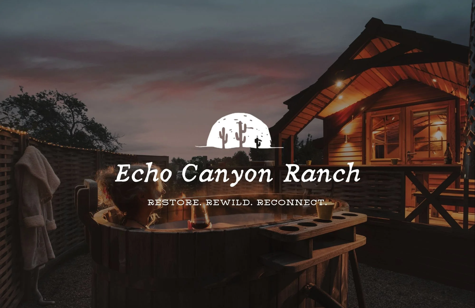 Nighttime scene at Echo Canyon Ranch showing a woman relaxing in a hot tub on a wooden deck, with a house illuminated by warm lights in the background, and a sunset sky with dark clouds.