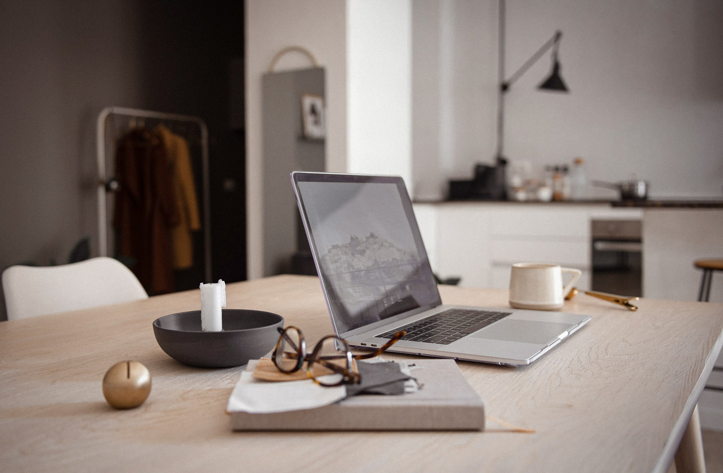 A laptop on a wooden table with a notebook, glasses, a candle in a black bowl, and a ceramic mug, in a modern kitchen or dining area.