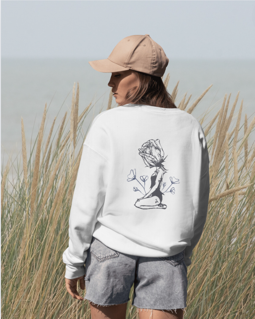 A woman with long brown hair wearing a beige baseball cap, white sweatshirt with a floral design on the back, and gray denim shorts stands in a field of tall beige grass near the water.