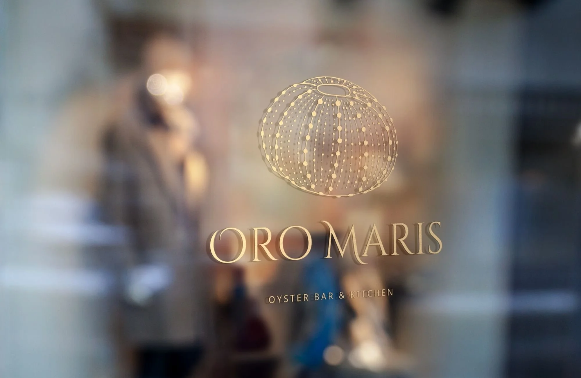 Gold foil logo for a high-end seafood restaurant, featuring a sea urchin-inspired emblem on a glass window with a blurred interior background.